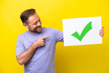 Middle age caucasian man isolated on yellow background holding a placard with text Green check mark icon and  pointing it