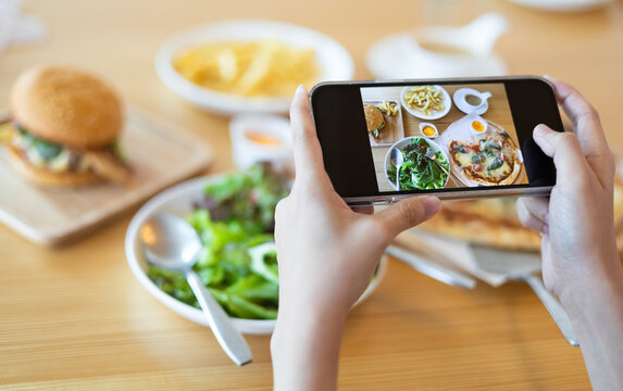 Close up of young woman eating hamburger, pizza, French fries and salad at restaurant and holding smartphone for take her food photo. Enjoy eating concept. Take a photos concept.