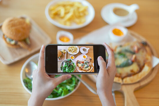 Woman captured her food with smart phone with feeling happy and enjoy. Enjoy eating, Take a photos concept. Hand holding smartphone against close up of pizza, french fries and salad at restaurant.