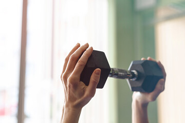 Side view of fit adult slim woman lifting weight in fitness box. Close-up on the arm with muscles in front of the window. Workout at home or intdoors. Only hands