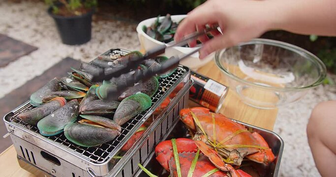Woman roasting fresh Green clam Shell mussels with tongs and crab grilling on a hot camping stove, cooking seafood party outdoor