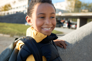 Adorable male child smiling at camera over blurred background of city landscape , going to school in sunny morning, carrying black satchel on shoulders, dressed in yellow hoodie and puffer vest