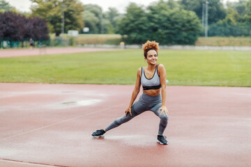 Fototapeta premium A happy, slim runner stretching her legs and warming up while standing in the stadium on a rainy day and listening music on the headphones. A runner in the stadium is warming up.