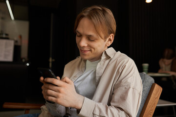 Attractive young Caucasian guy chatting with his girlfriend or reading comments on social networks, using smartphone, looking at screen, typing message with cute smile, sitting in chair at cozy cafe