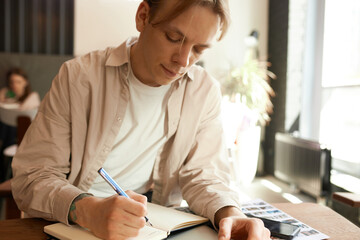 Close-up portrait of smart creative blond man in beige shirt writing in notebook with fountain pen...