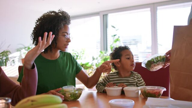 Multiracial family sitting at table and having take away food with children at home.
