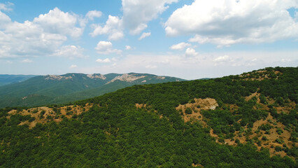 Obraz premium Aerial view of the grey mountain ranges and mountain slopes covered by green trees and shrubs against the blue cloudy sky in summer day. Shot. Beautiful mountain scenery