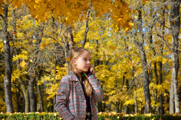 beautiful girl in a plaid coat talking on the phone in the autumn park. the child enjoys the warm rays and the beauty of autumn nature