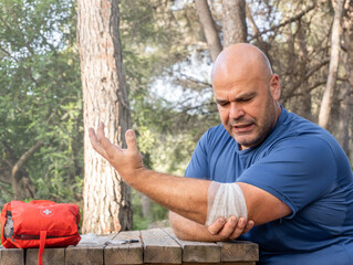 Male hiker sitting next to a red first aid kit, putting a bandage on his elbow. 