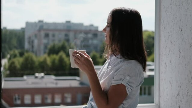 Pretty young woman on balcony looking out over city drinking a cup of tea or coffee