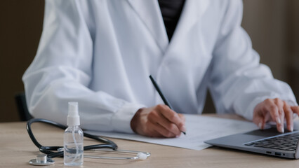 Close up unrecognizable female hand worker doctor practitioner writing prescription noticing documents at desk with medical equipment therapist woman handwriting notes typing use computer at hospital