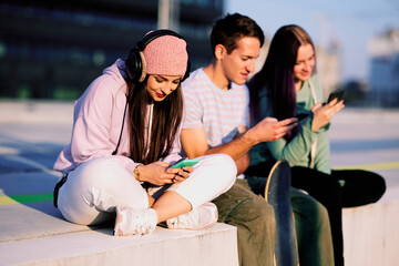 Three teenage friends are sitting in the urban exterior and hanging on their phones.