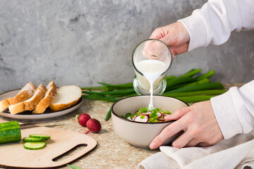 Female hands pour kefir into a bowl with chopped vegetables for a traditional Russian cold soup