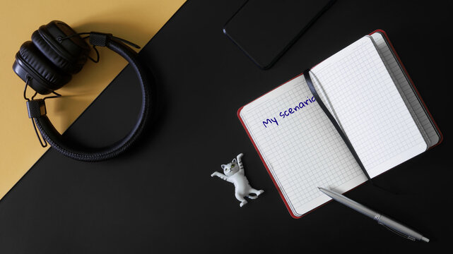 Headphones, smartphone, toy mascot cat, pen and notepad with scenario inscription on black and yellow background. Concept of writing screenplay, the work of a screenwriter and screenwriting.