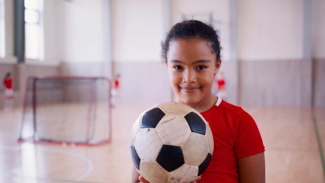 Small multiracial girl standing with ball indoors in gym class, physical education concept