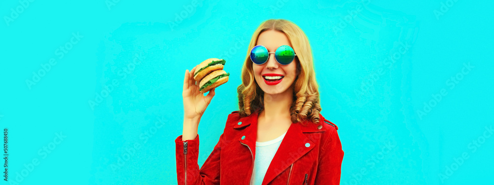 Wall mural portrait of stylish happy smiling young woman with tasty big burger fast food on blue background, bl