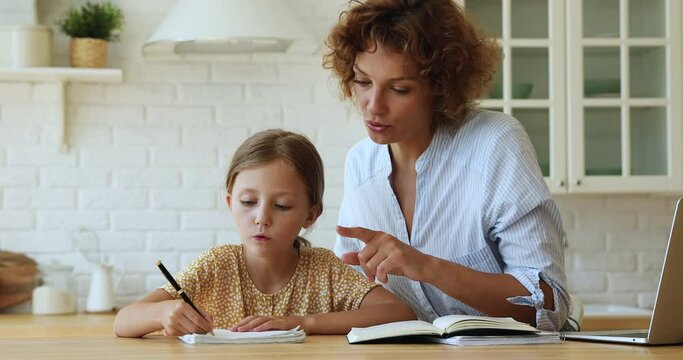 Happy young mother or female tutor educating little kid girl at home. Focused small child daughter preparing homework studying with caring mommy, handwriting notes in copybook, learning concept.