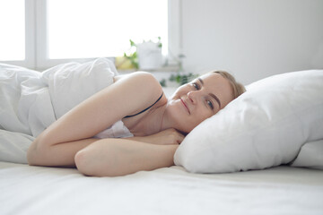 Positive blonde 30s woman with pillow lying in bed and smiling to camera at home. 