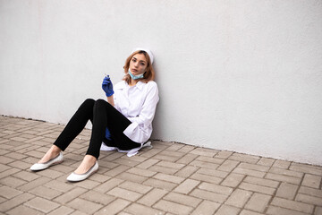 Dirty nurse with ash on face sitting outside hospital infirmary smoking cigarette after hard working day or surgery. Tired exhausted doctor woman dressed white medical gown have a rest due to stress
