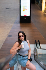 CHICA JOVEN SONRIENTE SUBIENDO ESCALERAS MECANICAS CON BATIDO