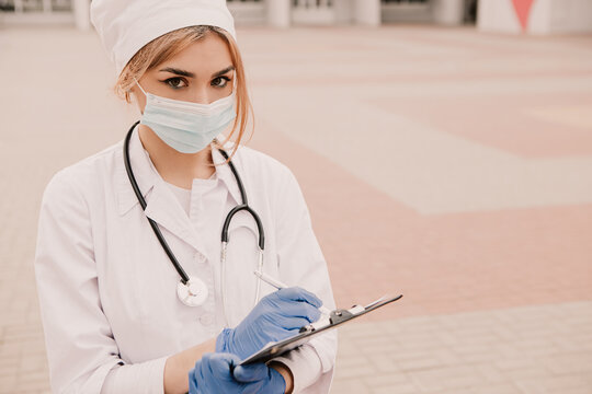 Young Female Nurse Standing Outside Hospital Infirmary Writing Patient Illness History In Journal. Doctor Woman Dressed White Medical Gown, Rubber Blue Gloves, Stethescope And Face Mask Fill Paper Doc