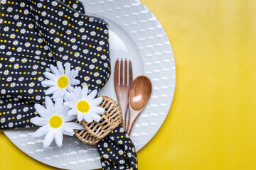 Summer dining concept with white plate, wicker napkin ring, daisies and wood fork and spoon on...