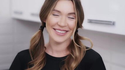 Close-up indoor portrait of a charming and a bit shy dark blond woman. Pretty girl smiling when looking into the camera and touching her pigtails