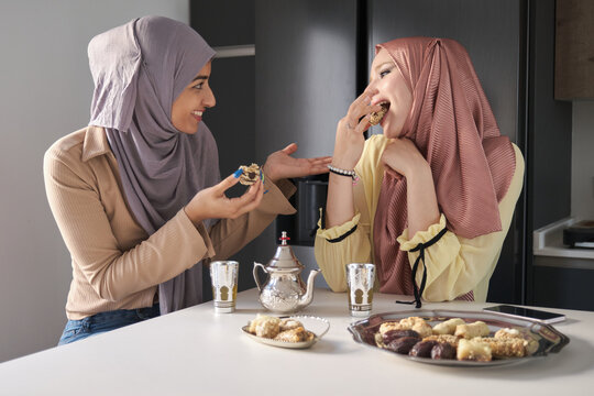 Two Young Muslim Women Talking, Drinking Tea And Eating Arabic Sweets At The Kitchen Table.