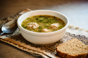 Homemade appetizing, delicious soup with meatballs, homemade bread, kitchen national napkin, and spoon on the kitchen dining table.