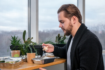 The businessman took a break and eats healthy food from a disposable dish. young handsome man smiles at the camera and holds a salad to eat. The guy is having lunch by the window