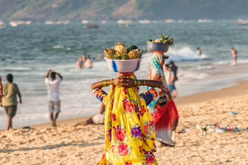 Unidentified Indian woman in colorful pink blue sari dress carrying fresh fruits in red basin at her head for selling at shoreline to tourists, CALANGUTE, GOA, INDIA - obrazy, fototapety, plakaty