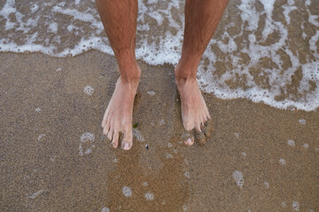 The guy stands on the sand barefoot. Legs close up. Sea. Ocean