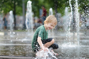 Little boy plays in the square between the water jets in the city fountain at sunny summer day. Active summer leisure for kids in city.