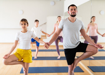 Fototapeta na wymiar Friendly sporty family of four practicing partner yoga in fitness studio. Teen girl with father standing on one leg in Eka Pada Pranamasana pose holding hands