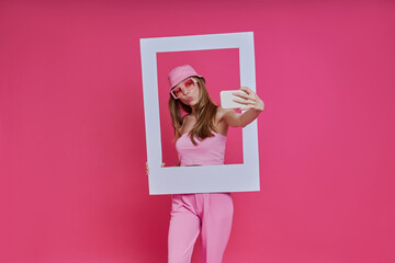 Playful young woman looking through a picture frame while standing against pink background