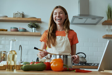 Happy young woman cooking soup and smiling while standing at the domestic kitchen