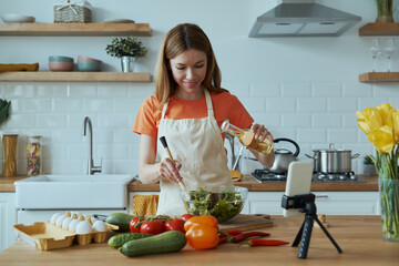 Beautiful young woman cooking and vlogging while standing at the domestic kitchen