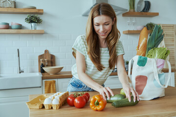 Beautiful young woman unpacking the bag with healthy food while standing at the domestic kitchen