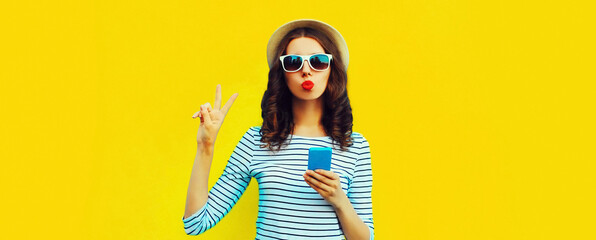 Portrait of happy young woman blowing her lips posing with smartphone wearing summer straw hat on...