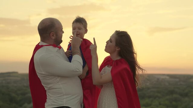 Friendly family concept, Happy family in superhero red capes are playing in park in sun. Mom dad kid dream together, family team. Father and son, mother, play superheroes at sunset. Happy childhood