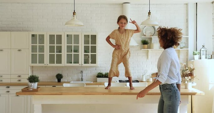 Happy young mother having fun with crazy emotional little kid daughter standing on table, dancing to favorite disco music in modern kitchen, celebrating freedom on weekend or moving into new dwelling.