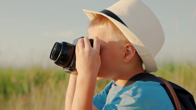 A cute little kid is holding vintage camera and taking pictures of nature. Happy boy dreams of becoming photographer. Kid plays in park in summer, spring. Child dreams of traveling, making discoveries
