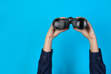 Woman holding modern binoculars on light blue background, closeup. Space for text