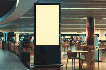 A blank vertical advertising billboard in front of a crowded mall food court with tables and...