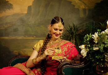 Indian bride dressed in Hindu red traditional wedding clothes sitting on sofa holding small gold box. - Powered by Adobe