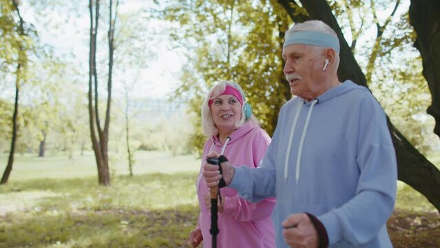 Cheerful senior elderly caucasian couple jogger grandparents training Nordic walking with ski trekking poles, running in summer park outdoor. Mature grandfather, grandmother hiking leisure sport hobby