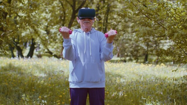 Modern old man exercising making dumbbell weight lifting exercises, practicing sport training in park wearing VR helmet to play simulation video game. Fitness for seniors. Active retired sportsman