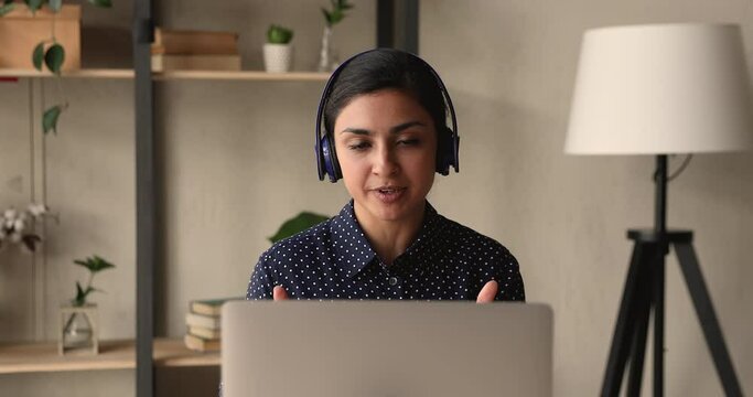 Indian woman sit at desk wear headphones looks at laptop screen talking pass job interview remotely, answers questions communicates to employer using video call application, modern tech usage concept