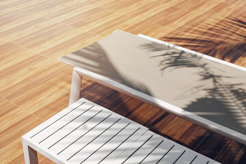 A sunbed with a wooden table under the shade on sunny day. Shadow of the tropical palm tree. Outdoor lounger for relaxation. Freshness and bright summertime vacation.