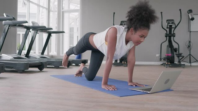 Fitness young black woman doing exercise yoga in gym. African american female loook at laptop computer lesson practicing Yaga plank pose on social online internet class. Healthy lifestyle concept.